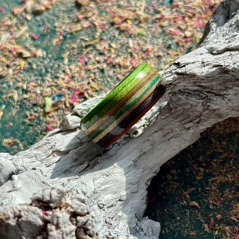 Green, purple and teal recycled skateboard wood ring resting on weathered wood, showing the full-width layered maple band.