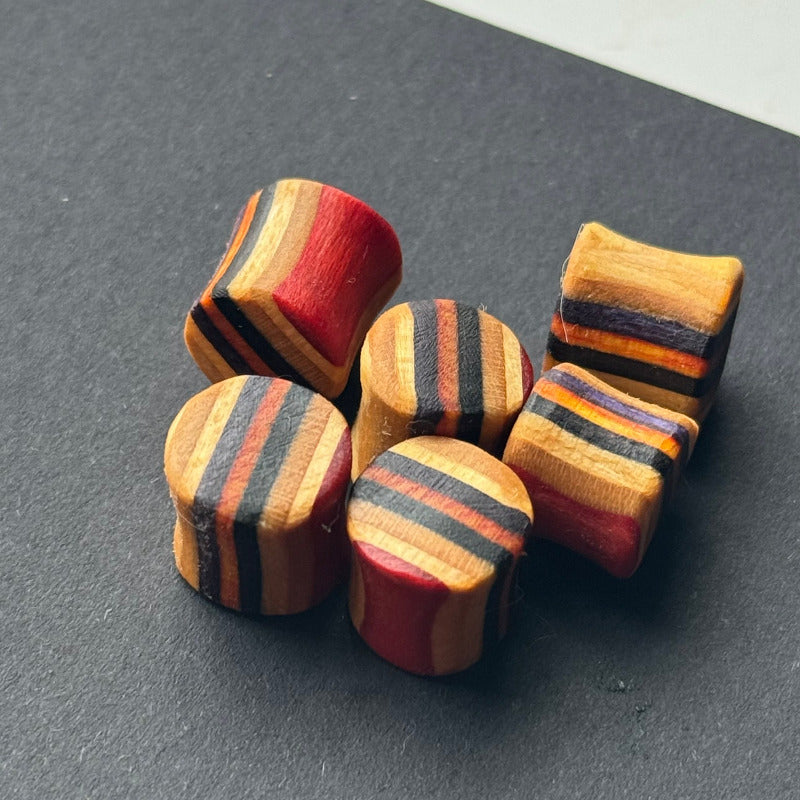 A group of 10mm wooden ear plugs made from recycled skateboard wood, featuring bold striped layers in red, black, orange, and natural tones, displayed on a dark surface in natural light.