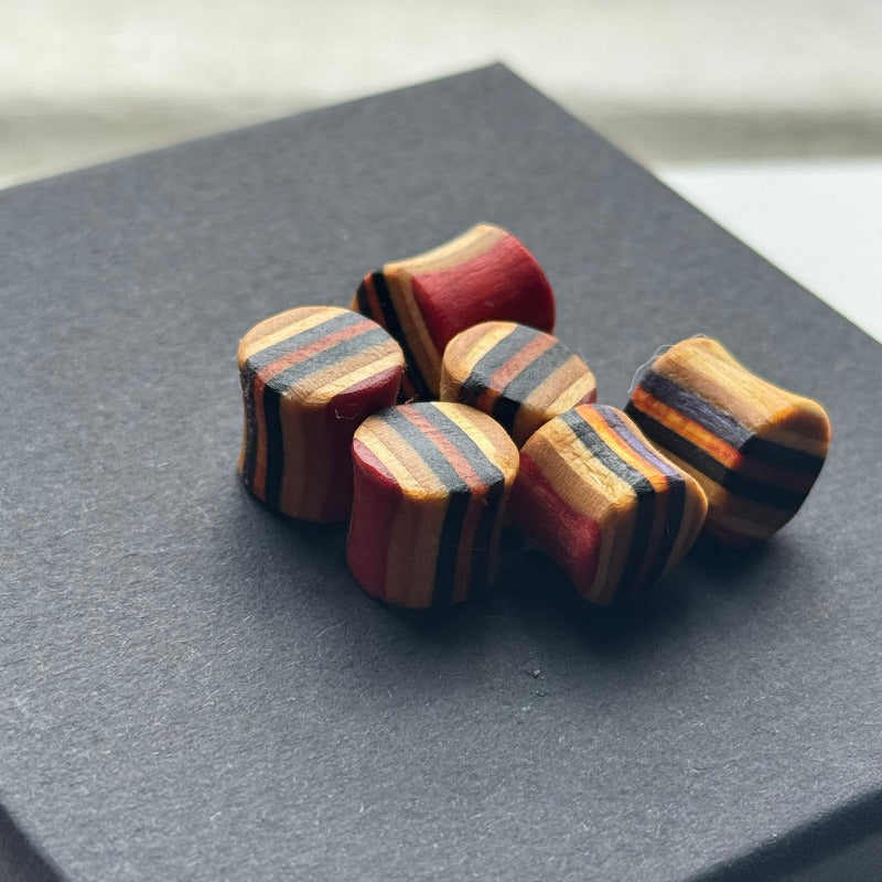 A group of 10mm wooden ear plugs made from recycled skateboard decks, showing layered stripes in red, orange, black, and natural wood tones, displayed on a dark box in soft daylight.